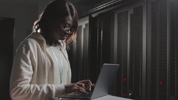 Pas-up mladého afroamerického ženského programátora s brýlemi, s použitím přenosného počítače, psaní v temné serverovně - Záběry, video