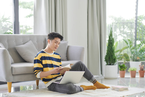 Aantrekkelijke gelukkige slimme jongeman zittend op een vloer in de woonkamer, werkend aan; laptop computer - Foto, afbeelding