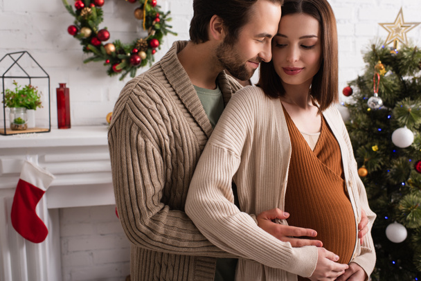 homme joyeux embrassant femme enceinte près flou arbre de Noël - Photo, image