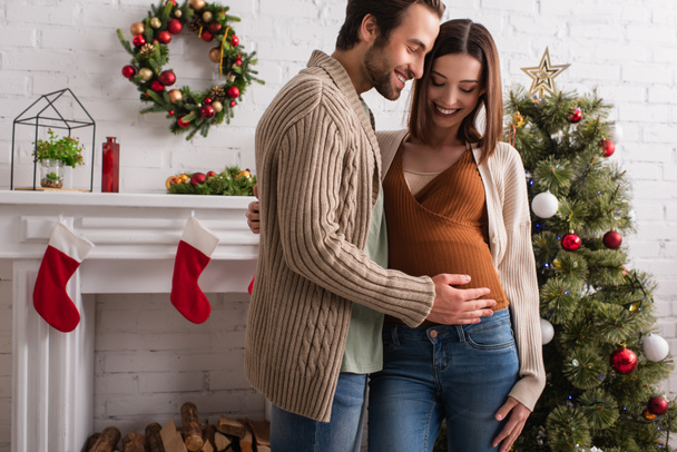 暖炉やクリスマスツリーの近くに笑顔の妻のおなかに触れる喜びの男 - 写真・画像