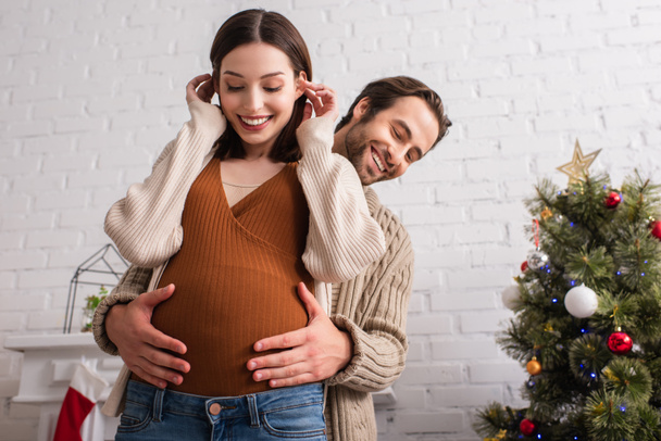 alegre hombre abrazando barriga de embarazada esposa cerca borrosa árbol de navidad - Foto, Imagen