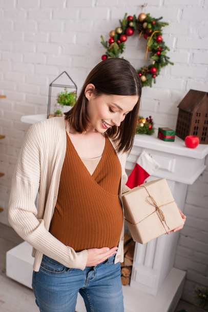 happy pregnant woman touching belly while holding christmas present near decorated fireplace - Photo, Image