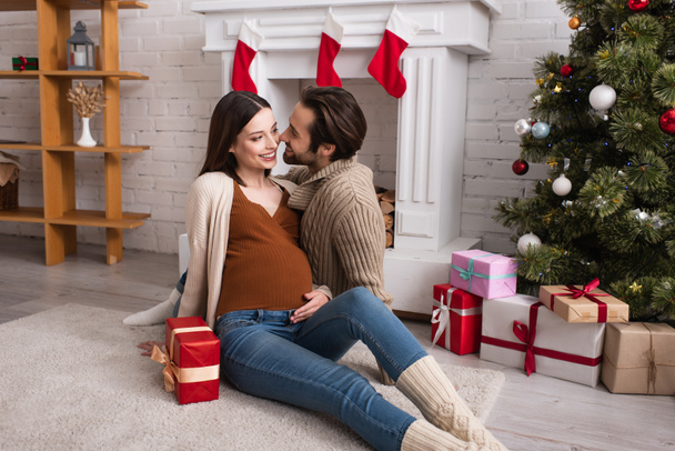 joyful pregnant woman with husband sitting on floor near decorated fireplace and presents under christmas tree - Photo, Image