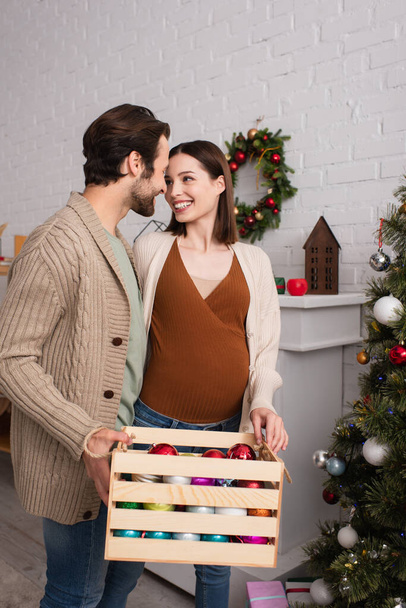 mujer embarazada feliz mirando marido sosteniendo caja de madera con bolas de Navidad - Foto, Imagen