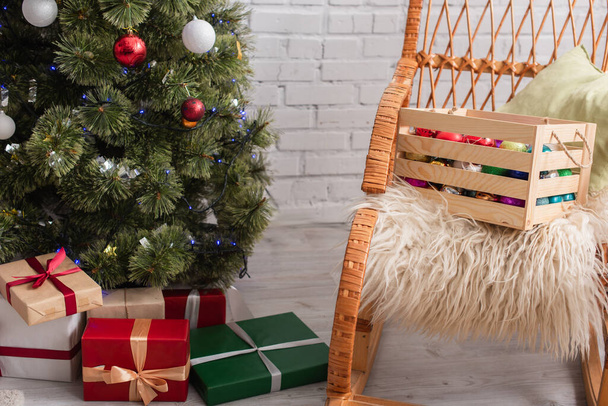wooden box with decorative baubles on wicker chair near presents under christmas tree - Photo, Image