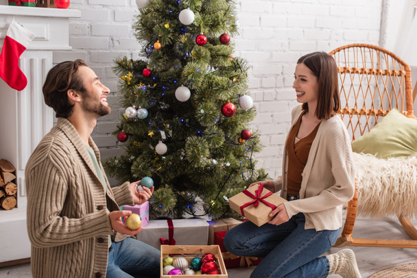 glückliches Paar mit Geschenk und Christbaumkugeln, die einander anlächeln, während sie auf dem Boden neben dem Weihnachtsbaum sitzen - Foto, Bild