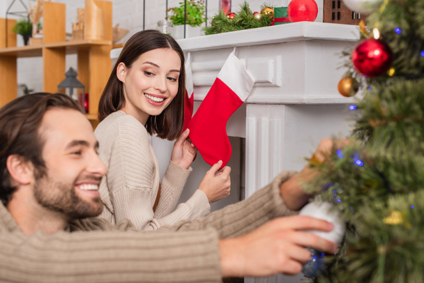 happy woman holding christmas stocking near fireplace and blurred husband decorating christmas tree - Photo, Image