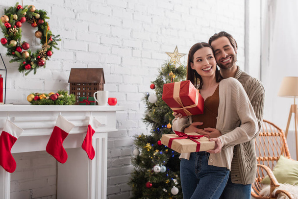 feliz hombre abrazando esposa sosteniendo cajas de regalo cerca del árbol de Navidad y chimenea decorada - Foto, Imagen