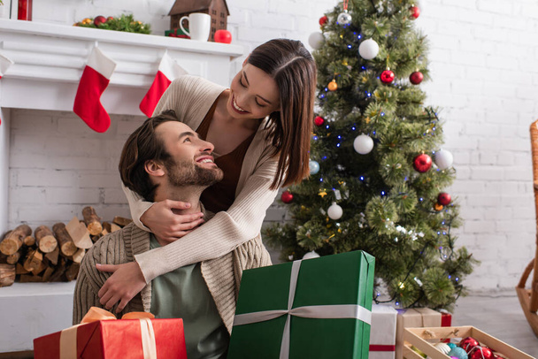 pleased woman embracing husband holding gift boxes near fireplace and christmas tree - Photo, Image