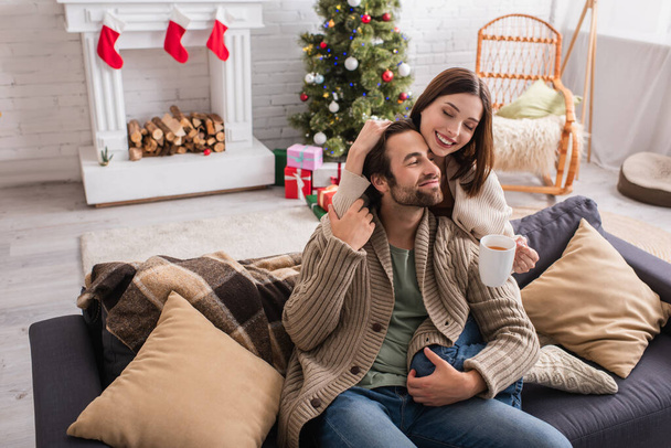 χαρούμενο ζευγάρι αγκαλιάζει τον καναπέ στο σαλόνι με χριστουγεννιάτικη διακόσμηση - Φωτογραφία, εικόνα