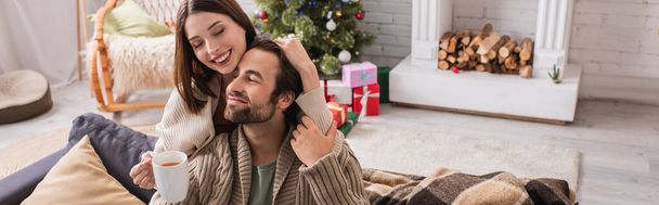 mulher alegre abraçando marido segurando xícara de chá na sala de estar festiva, banner - Foto, Imagem