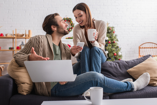 usmívající se žena s šálkem čaje ukazující smartphone manželovi ukazující na notebook v obývacím pokoji - Fotografie, Obrázek