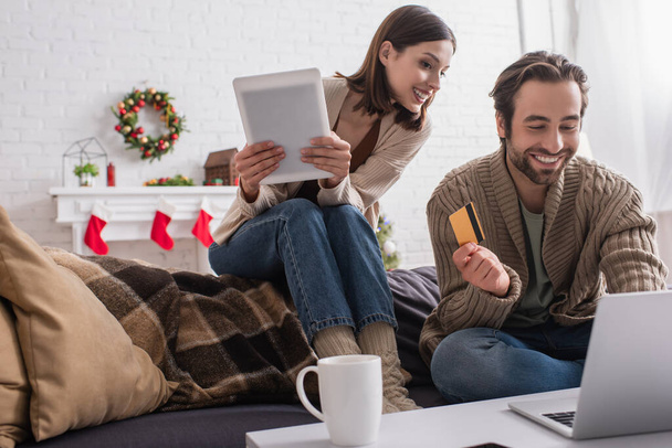 веселая пара с кредитной картой и цифровой планшет возле ноутбука в гостиной с праздничным оформлением - Фото, изображение