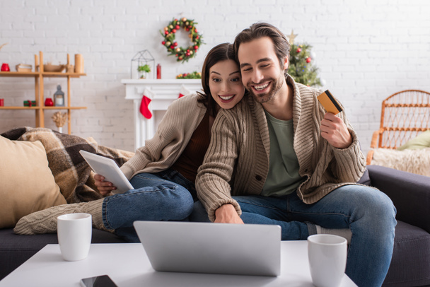 Dijital tabletli mutlu bir kadın ve dekore edilmiş oturma odasında dizüstü bilgisayara bakan kredi kartı olan bir adam. - Fotoğraf, Görsel