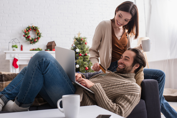 щасливий чоловік друкує на ноутбуці біля дружини з кредитною карткою у вітальні з різдвяним прикрасою
 - Фото, зображення