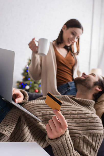 man with laptop holding credit card near blurred wife in living room - Photo, Image