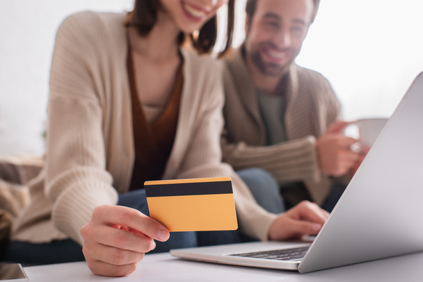 θολή γυναίκα κρατώντας πιστωτική κάρτα κοντά στο laptop και χαμογελαστός σύζυγος στο σπίτι - Φωτογραφία, εικόνα