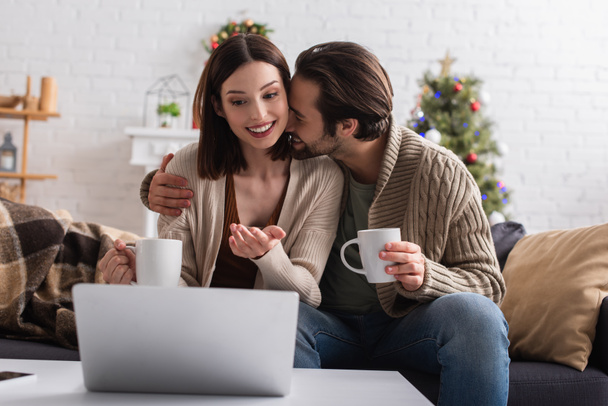 smiling woman pointing at blurred laptop near husband hugging her on sofa at home - Photo, Image