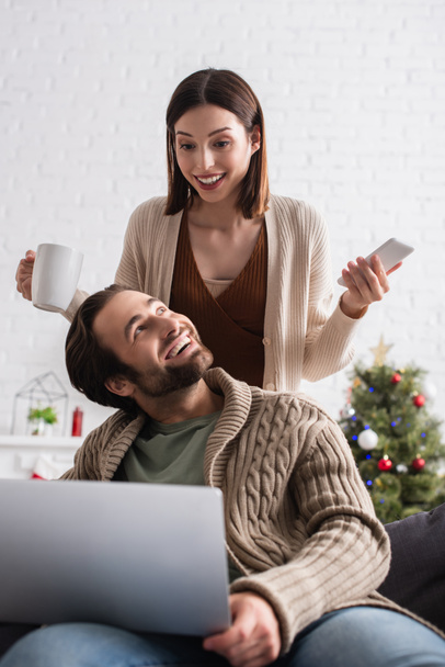 positive woman holding smartphone and cup of tea near cheerful husband and laptop - Photo, Image