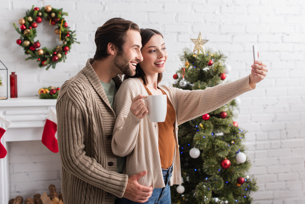 iloinen nainen tilalla kuppi teetä ottaen selfie miehensä kanssa lähellä hämärtynyt joulukuusi - Valokuva, kuva