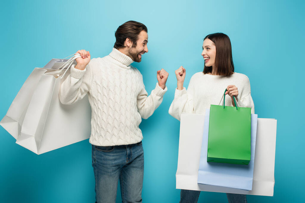 cheerful couple with shopping bags showing success gesture on blue - Photo, Image
