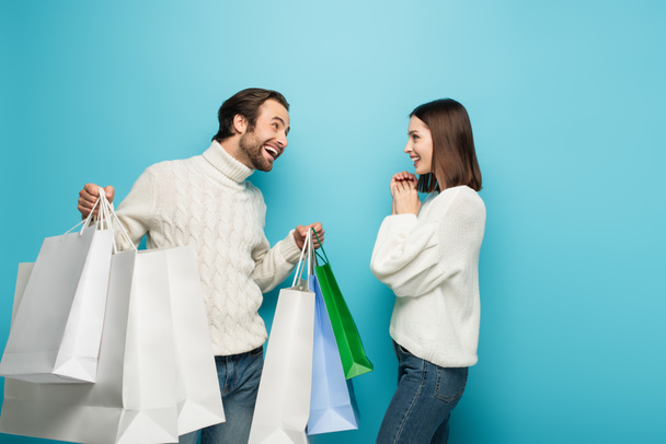 alegre hombre sosteniendo bolsas de compras cerca de mujer sorprendida en azul - Foto, imagen
