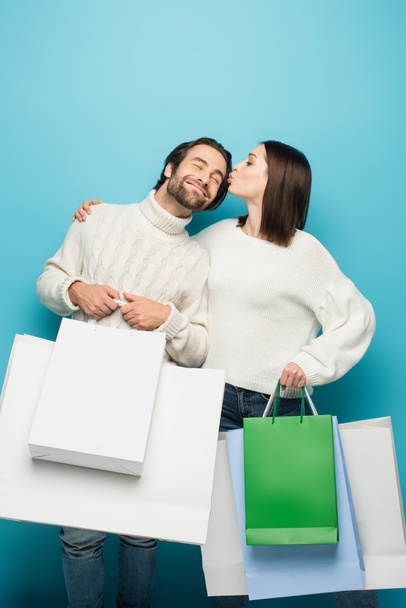 woman holding shopping bags and kissing pleased man with purchases on blue - Photo, Image