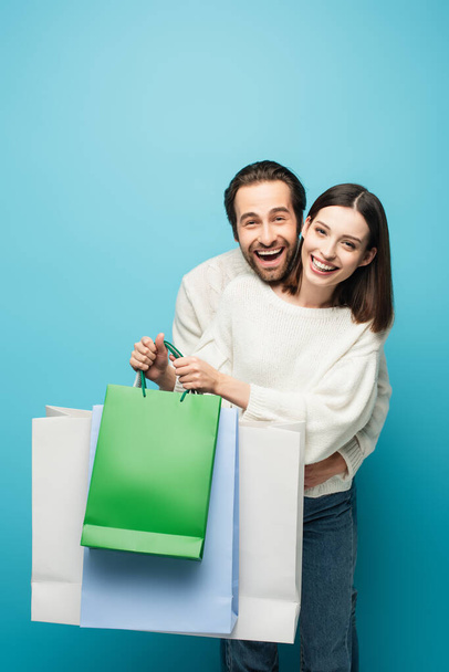 веселий чоловік обіймає жінку, тримаючи сумки для покупок ізольовані на синьому
 - Фото, зображення