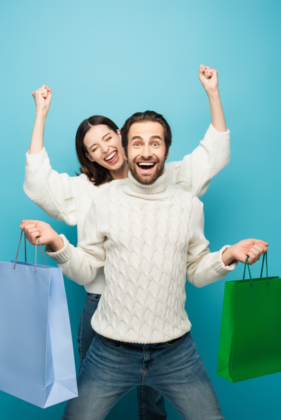astonished man holding shopping bags near woman showing yeah gesture isolated on blue - Photo, Image