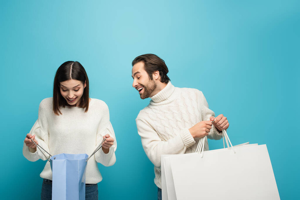 amazed woman looking into shopping bag near happy man with purchases on blue - Photo, Image