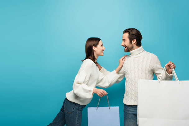 radosna para w zimowych swetrach trzymająca torby na zakupy i patrząca na siebie na niebiesko - Zdjęcie, obraz
