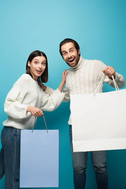 pareja asombrada con bolsas de compras mirando a la cámara en azul - Foto, Imagen