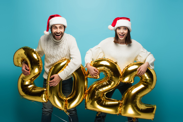 Aufgeregtes Paar in Weihnachtsmützen mit goldenen Luftballons in Form von 2022 auf blauem Grund - Foto, Bild