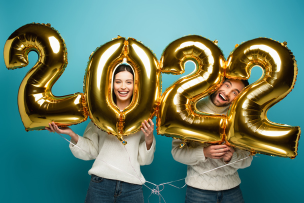 χαρούμενο ζευγάρι κοιτάζοντας κάμερα κοντά σε χρυσά 2022 μπαλόνια σε μπλε - Φωτογραφία, εικόνα