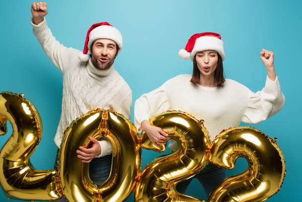 Aufgeregtes Paar in Weihnachtsmützen mit goldenen Luftballons in Form von 2022 und Siegergeste auf blauem Grund - Foto, Bild
