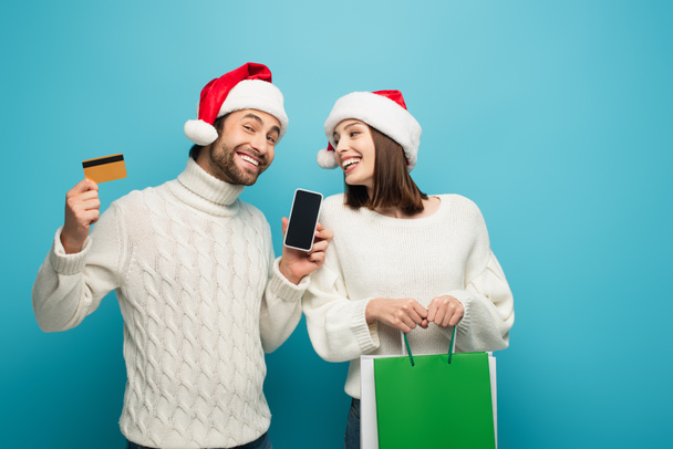 homme heureux dans santa chapeau tenant smartphone et carte de crédit près de la femme avec des achats sur bleu - Photo, image