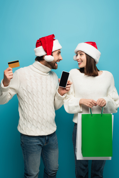 couple joyeux avec sacs à provisions, carte de crédit et smartphone se regardant sur bleu - Photo, image