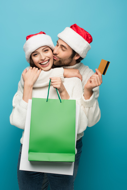 Mann mit Weihnachtsmannmütze küsst glückliche Frau mit Kreditkarte und Einkaufstaschen auf blauem Grund - Foto, Bild