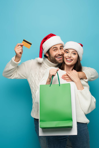 счастливый мужчина в шляпе Санты показывает кредитку и обнимает женщину с пакетами покупок на голубом - Фото, изображение