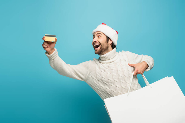 ενθουσιασμένος άνθρωπος με τσάντες ψώνια που δείχνουν πιστωτική κάρτα στο μπλε - Φωτογραφία, εικόνα