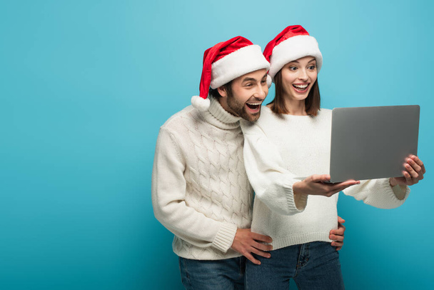 έκπληκτο ζευγάρι με ζεστά πουλόβερ και καπέλα santa κοιτάζοντας το φορητό υπολογιστή σε μπλε - Φωτογραφία, εικόνα