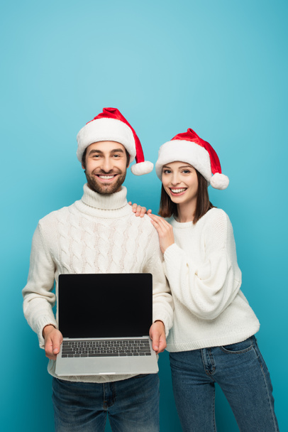 glücklicher Mann mit Weihnachtsmannmütze, Laptop mit leerem Bildschirm neben lächelnder Frau auf blauem Grund - Foto, Bild