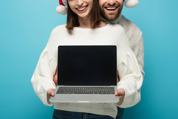 partial view of smiling couple in santa hats holding laptop with blank screen isolated on blue - Photo, Image