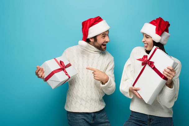 amazed man in santa hat pointing at gift box near woman sticking out tongue on blue - Photo, Image