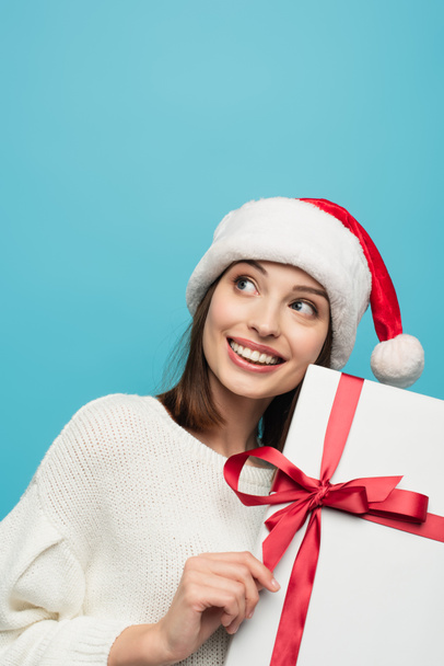 fröhliche Frau mit Weihnachtsmütze schaut weg, während sie Weihnachtsgeschenk isoliert auf blauem Grund hält - Foto, Bild