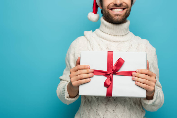 abgeschnittene Ansicht eines lächelnden Mannes mit Weihnachtsmütze und weißem Pullover, der Geschenke isoliert auf blauem Grund hält - Foto, Bild