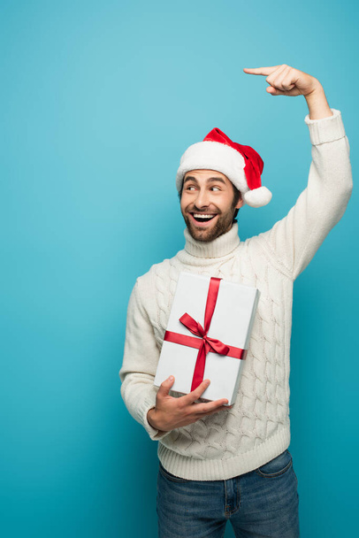 веселий чоловік у капелюсі Санта дивиться вбік і вказує пальцем, тримаючи подарунок ізольовано на синьому
 - Фото, зображення
