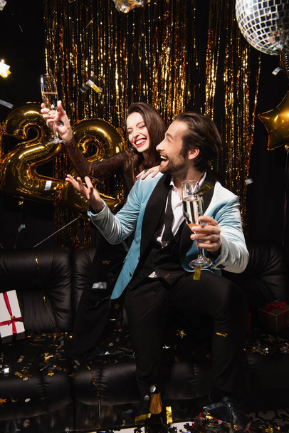 fröhliches Paar mit Champagner feiert Neujahr auf Sofa neben glänzender Dekoration auf Schwarz - Foto, Bild