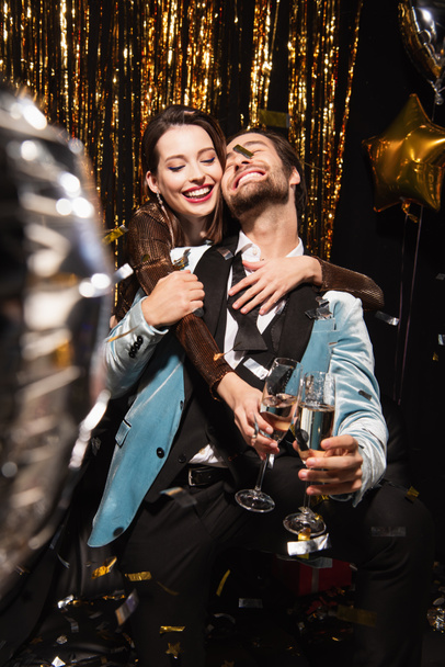 joyful and stylish couple clinking champagne glasses near sparkling christmas decoration on black - Photo, Image