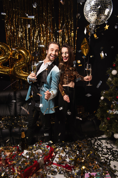veselý a elegantní pár s jiskry slaví Vánoce v blízkosti lesklé dekorace na černé - Fotografie, Obrázek